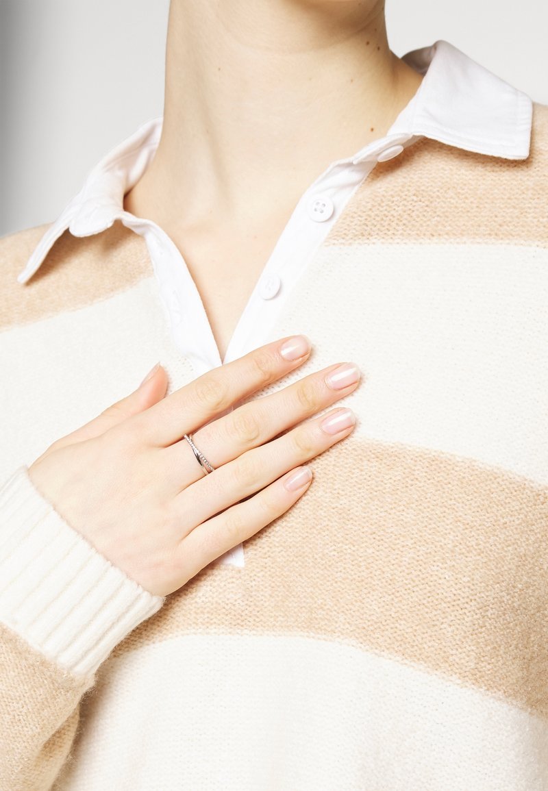 Main avec une bague en argent posée sur la poitrine, portant un pull beige et blanc rayé sur une chemise blanche à col.