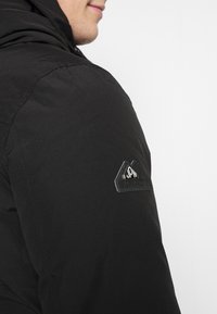 Černá bunda s hladkým povrchem, kapucí a trojúhelníkovou nášivkou na rukávu s názvem značky "Superdry."