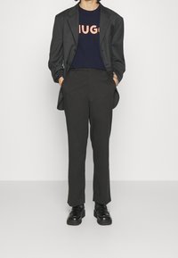 Hiilibliseri tummansinisellä T-paidalla, jossa on rohkea korallinvärinen "HUGO" -teksti, yhdistettynä tummiin housuihin ja mustiin kenkihin, joissa on paksu pohja.