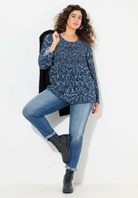 Tunika con stampa animale blu e vita arricciata, abbinata a jeans azzurri e stivali neri con lacci alla caviglia. Giacca nera appoggiata sul braccio.