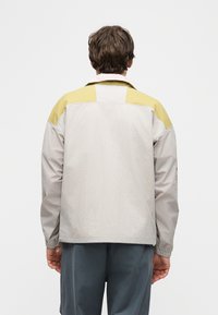 Άνδρας με την πλάτη στραμμένη, φοράει ένα ανοιχτό γκρι μπουφάν με κίτρινες επιφάνειες στους ώμους και σκούρα γκρι παντελόνια, με φόντο ένα απλό λευκό φόντο.