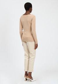 Beige Pullover mit langen Ärmeln und rundem Ausschnitt, kombiniert mit cremefarbenen Cuffed-Hosen und weißen High-Heels. Glatte Textur, figurschnittdesign.