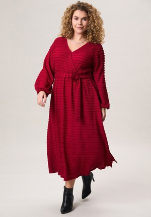 Vrouw met krullend haar, gekleed in een gestructureerde rode wikkeljurk en zwarte enkellaarzen, lopend naar voren op een lichtgrijze achtergrond.