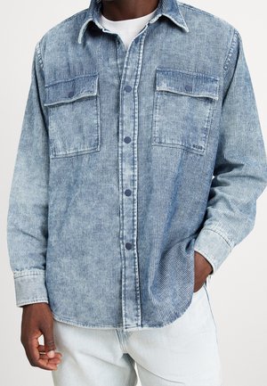 Man draagt een vervaagde blauwe ribfluwelen blouse met borstzakken en lichtblauwe spijkerbroek, één hand in de zak.