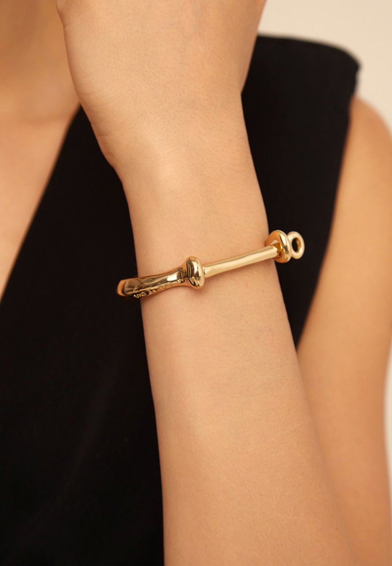 Pulsera rígida de metal dorado con un diseño liso y curvado que presenta dos extremos redondeados distintivos y una superficie texturizada.