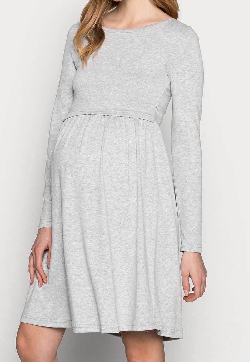 Femme portant une robe de maternité gris clair à manches longues, taille empire et jupe longueur genou sur un fond uni.