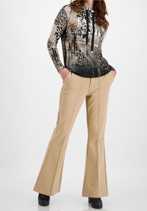 Woman wearing beige flared trousers, black platform shoes, and a long-sleeve animal print top with black drawstrings, hands in pockets.