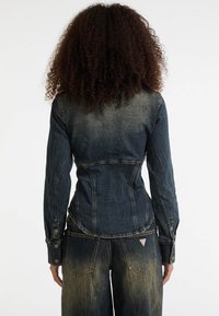Denimjack in donkerblauw, aansluitend ontwerp met lange mouwen, knoopaccenten en gestikte details. Achteraanzicht toont een lichte vervaging van de stof.