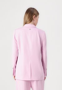 Blazer entallado de color rosa claro con cierre de un solo botón, abertura en la parte posterior y mangas largas. Textura suave con un brillo sutil.