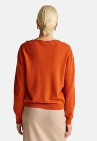 Pomarańczowy sweter na drutach o luźnym kroju, okrągłym dekolcie i ściągaczach przy mankietach, w zestawieniu z beżową spódnicą. Gładka tekstura, miękki materiał.