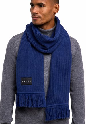 Écharpe tricotée bleue avec un motif texturé, aux extrémités frangées, et une étiquette noire avec du texte blanc, portée sur un pull gris.
