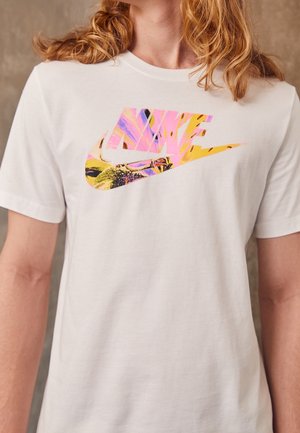 T-shirt blanc avec un logo Nike coloré et abstrait porté par une personne aux longs cheveux roux ondulés, visible des épaules à la partie supérieure de la poitrine.