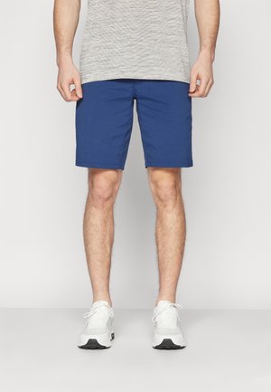 Man met een lichtgrijs T-shirt, blauwe knielange korte broek en witte sneakers, staand tegen een effen lichte achtergrond.