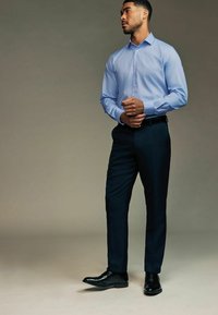 Hellblaues Hemd mit Knöpfen, kombiniert mit dunkelblauen Hosen. Modell trägt schwarze Lederschuhe, steht mit verschränkten Armen vor neutralem Hintergrund.