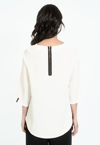 Blusa bianca con maniche a tre quarti, scollatura rotonda e dettaglio zip nera sul retro. Tessuto morbido e liscio con una vestibilità ampia.