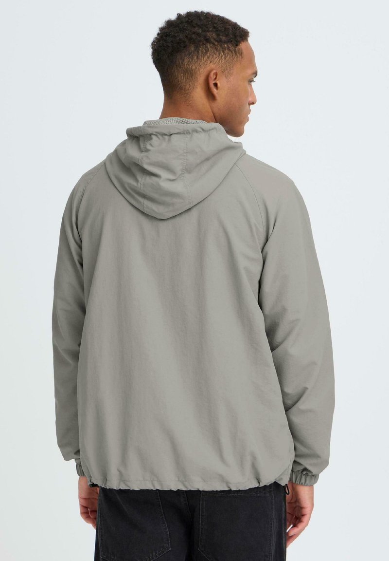 Jeune homme vu de dos portant une veste à capuche gris clair avec des poignets élastiques et un pantalon noir, sur un fond blanc uni.