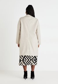 Szary wełniany płaszcz o prostym kroju i otwartym froncie, noszony na spódnicy w czarno-biały geometryczny wzór oraz czarnych botkach na obcasie.