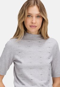 Jasnoszara, krótkosleeve'owa, dzianinowa bluzka z wysokim kołnierzem, zdobiona metalicznymi elementami ułożonymi w poziome paski na materiale.