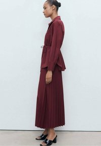 Gonna midi plissé color bordeaux abbinata a una blusa con cintura coordinata. L'outfit presenta una vestibilità rilassata e sandali con tacco neri.