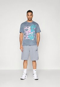 Harmaa Nike-t-paita, jossa iso kuviointi turkoosilla ja vaaleanpunaisella, yhdistetty vaaleanharmaisiin shortseihin ja valkoisiin Nike-tennareihin.