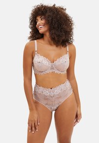 Femme souriante aux cheveux bouclés portant un soutien-gorge en dentelle rose pâle assorti à une culotte taille haute, regardant vers sa gauche sur fond blanc.