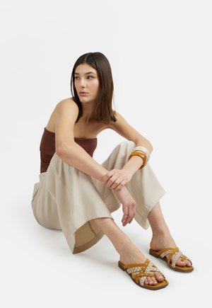 Giovane donna seduta sul pavimento con top marrone senza spalline, pantaloni beige a gambe larghe, braccialetti in ambra e sandali piatti intrecciati color cuoio.