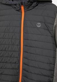 Kurtka puchowa bez rękawów w kolorze czarnym z kapturem, z pomarańczowym zamkiem błyskawicznym i szarymi rękawami. Posiada okrągłe logo na klatce piersiowej oraz gładką teksturę.