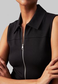 Nahbild av en modell i en ärmlös svart zip-up topp, med korslagda armar, som visar en polerad look mot en neutral bakgrund.