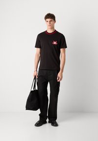 Black short-sleeve T-shirt with red ribbed collar and a graphic print. Paired with black cargo pants and carrying a black tote bag.