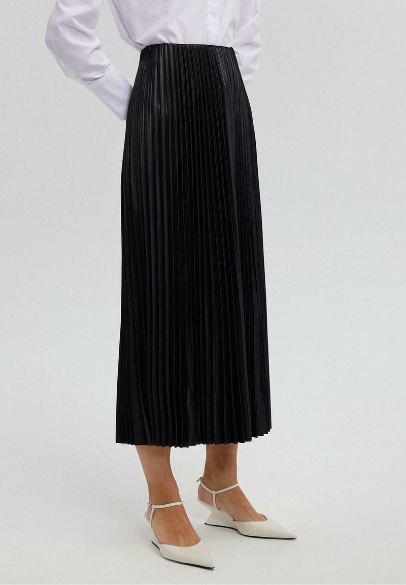 Jupe midi plissée noire à taille haute, en tissu brillant, associée à des escarpins blancs à bout pointu et une chemise boutonnée.