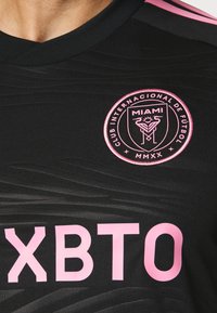 Musta urheilupaita, jossa on teksturoitu kuvio ja pyöreä, vaaleanpunaisella tekstillä oleva "MIAMI"-logo sekä voimakkaasti korostettu "XBTO"-teksti vaaleanpunaisena.