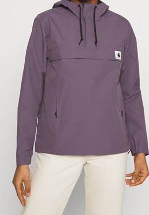 Persoon met een paarse anorak met capuchon en zwarte trekkoorden, en lichtbeige broek, staand tegen een effen achtergrond.