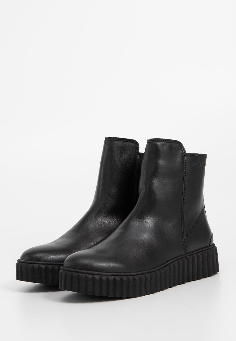 Ein Paar schwarze Leder-Stiefeletten mit dicken, gerippten Gummisohlen und seitlichen Nähten, auf einem weißen Hintergrund präsentiert.