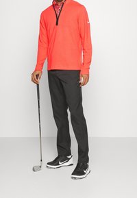 Homme en pantalons noirs et chaussures de golf Nike noir et blanc tenant un club de golf, portant un pullover rouge à quart de zip sur une chemise à motifs rouges.