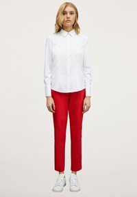 Camisa blanca de manga larga con cuello, combinada con pantalones rojos ajustados y zapatillas blancas. El conjunto presenta un diseño limpio y minimalista.