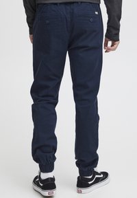 Pantalon de jogging bleu marine avec des poignets élastiques, deux poches arrière et un léger éclat. Porté avec des baskets noires et blanches.