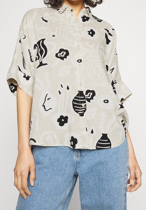 Vrouw met een beige overhemd met knopen, bedrukt met zwart-witte abstracte bloemen en vazen, gecombineerd met lichtblauwe spijkerbroeken.