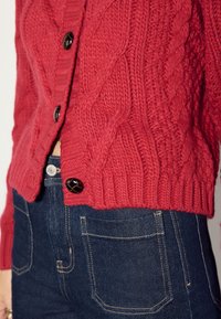 Red knitted cardigan with a textured cable pattern and black buttons, paired with dark denim jeans featuring contrasting stitching.