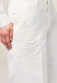 Pantalones de algodón blancos con bordado floral texturizado en el lado izquierdo. Incluyen bolsillos tradicionales y un cierre de cremallera.