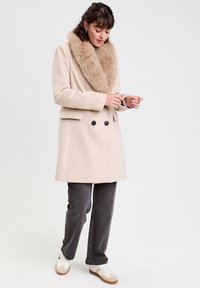 Manteau beige croisé avec un col en fausse fourrure douce et brun clair, associé à un jean gris et des baskets blanches. Design lisse et ajusté.