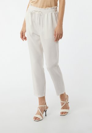 Kobieta ubrana w białe spodnie z wysokim stanem i sznurkiem oraz beżowe sandały na wysokim obcasie na jednolitym tle.