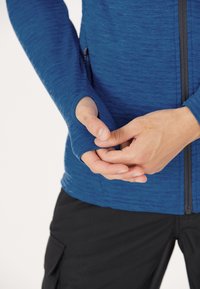 Långärmad blå träningsskjorta med strukturerad tyg, tumhål vid ärmsluten och full dragkedja fram; bärs över svarta byxor.