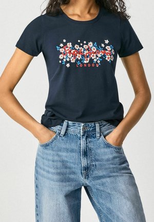 Mujer con camiseta gráfica azul marino con flores y texto "Pepe Jeans London", combinada con jeans de mezclilla azul claro, con las manos en los bolsillos.