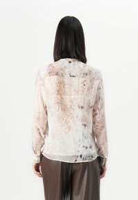 Frau mit geraden, dunklen Haaren trägt eine durchsichtige Bluse mit langen Ärmeln in hellbraunem und schwarzem gesprenkeltem Muster sowie eine dunkle weit geschnittene Hose, Rückansicht.
