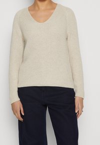 Strickpullover in Creme mit V-Ausschnitt, langen Raglanärmeln und strukturiertem Finish. Kombiniert mit dunkelblauen weiten Hosen.