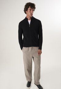 Zwarte zip-up jas met een hoge kraag, gecombineerd met lichtgrijze broek en zwarte sneakers. Glad textuur, ontspannen pasvorm en casual stijl.
