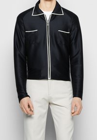 Man som bär en svart zip-up jacka med vit kant och två bröstfickor över vita byxor mot en neutral bakgrund.