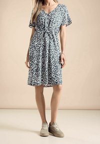 Donna che indossa un vestito corto a maniche corte, fino al ginocchio, con stampa di foglie verde scuro e sneakers beige, in piedi di fronte a uno sfondo beige uniforme.