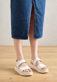 White platform sandals with woven texture, featuring adjustable straps and a buckle. Set against a denim skirt and wooden floor backdrop.