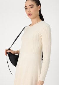 Abito a coste beige con maniche lunghe e scollatura rotonda, caratterizzato da una parte superiore testurizzata e una sezione inferiore liscia. Una borsa nera è tenuta in mano.